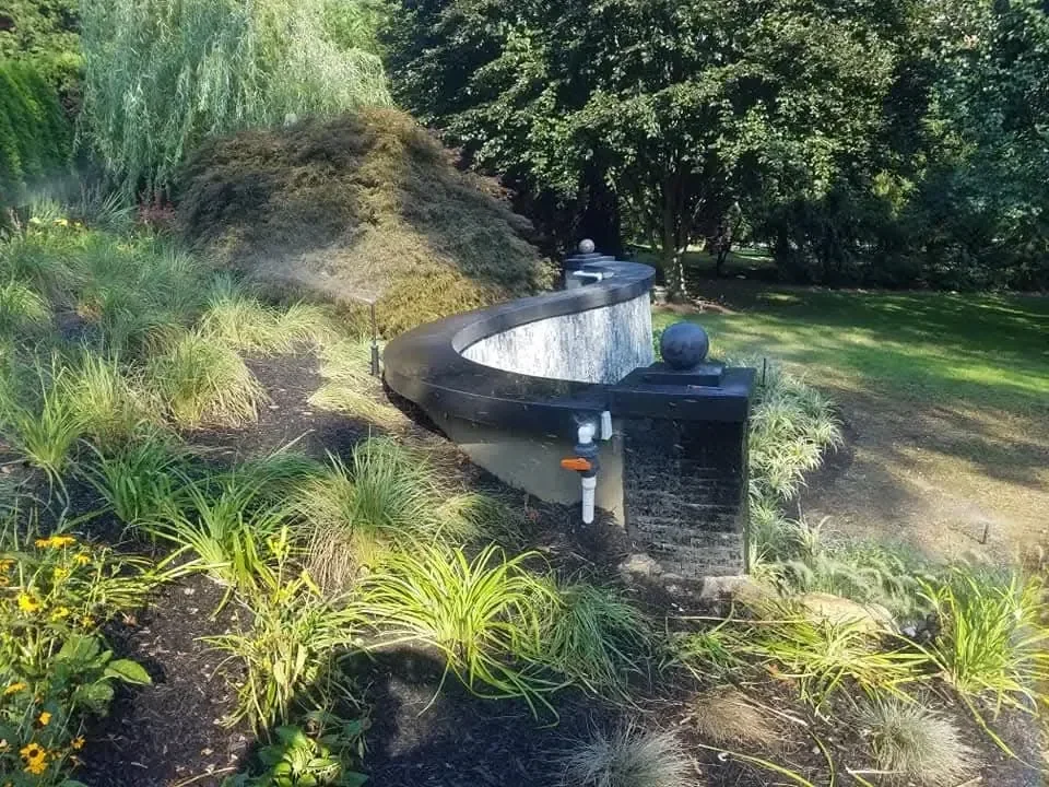 A curved, dark stone water feature is nestled among lush ornamental grasses and mature trees in a manicured garden setting.