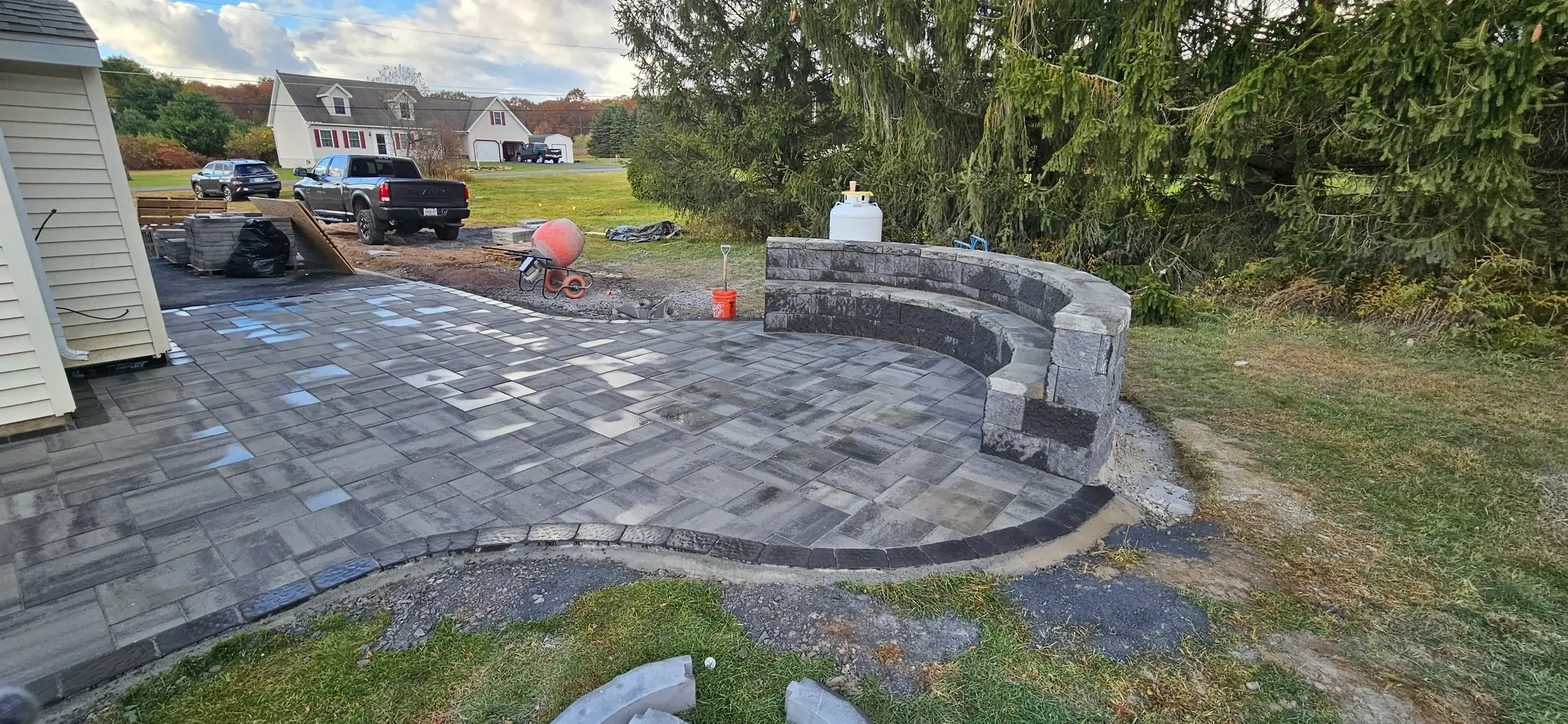 A newly installed gray paver patio featuring a curved, tiered stone seating wall is shown amidst ongoing residential hardscaping work.