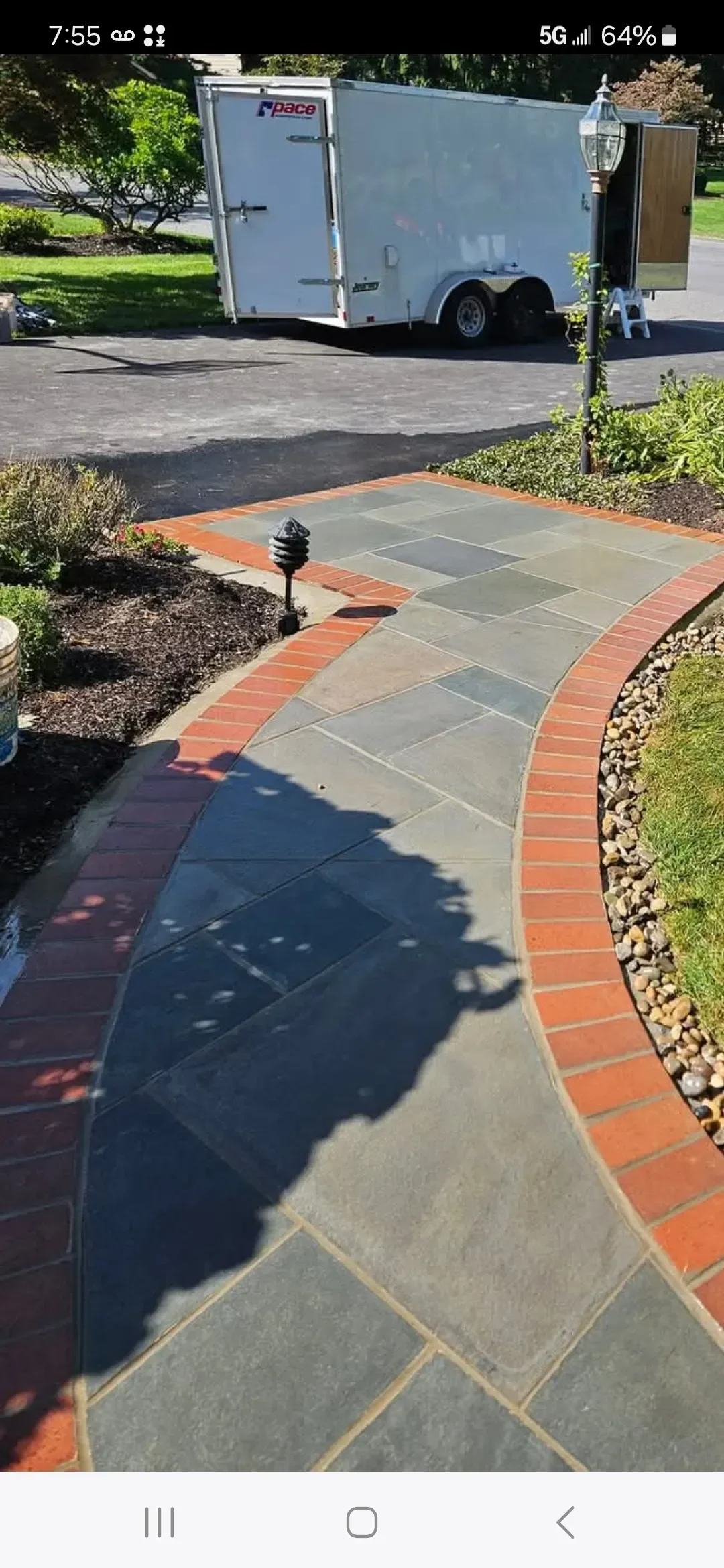 A newly installed curved walkway featuring gray stone pavers bordered by red brick is visible in a residential yard, with a white utility trailer parked on the adjacent asphalt driveway.