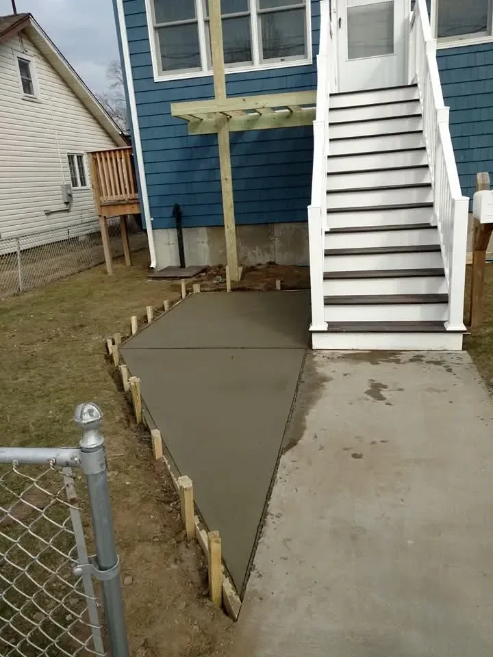 A freshly poured concrete walkway, still held by wooden forms, runs alongside a blue-sided house with a long white exterior staircase and an adjacent chain-link fence.