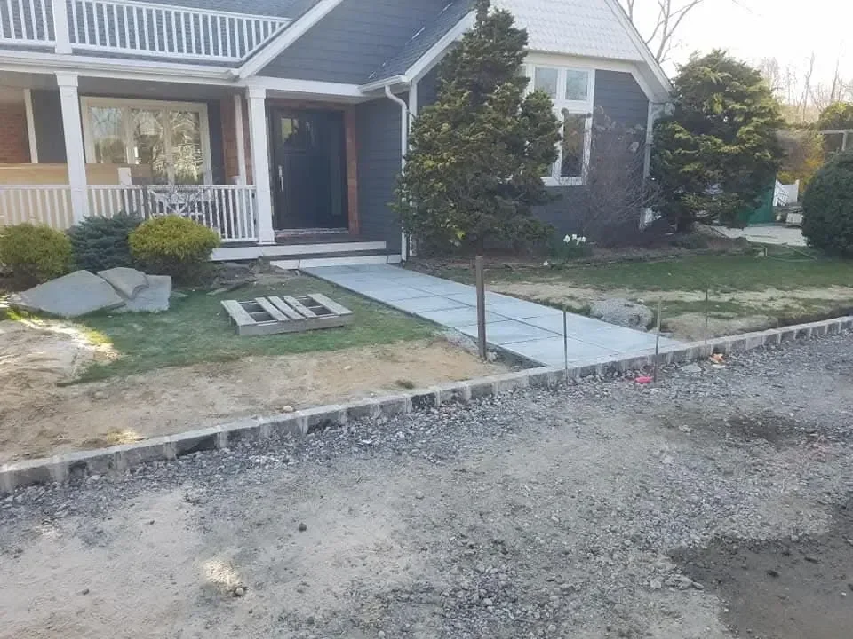 A new paved walkway and stone edging are being installed in the front yard of a dark-sided residential home.