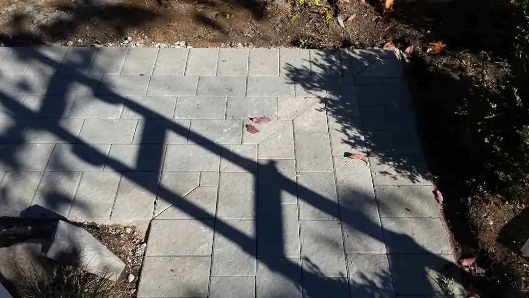 An overhead view shows newly installed gray rectangular pavers forming a walkway or patio bordered by dark soil and vegetation under strong diagonal shadows.