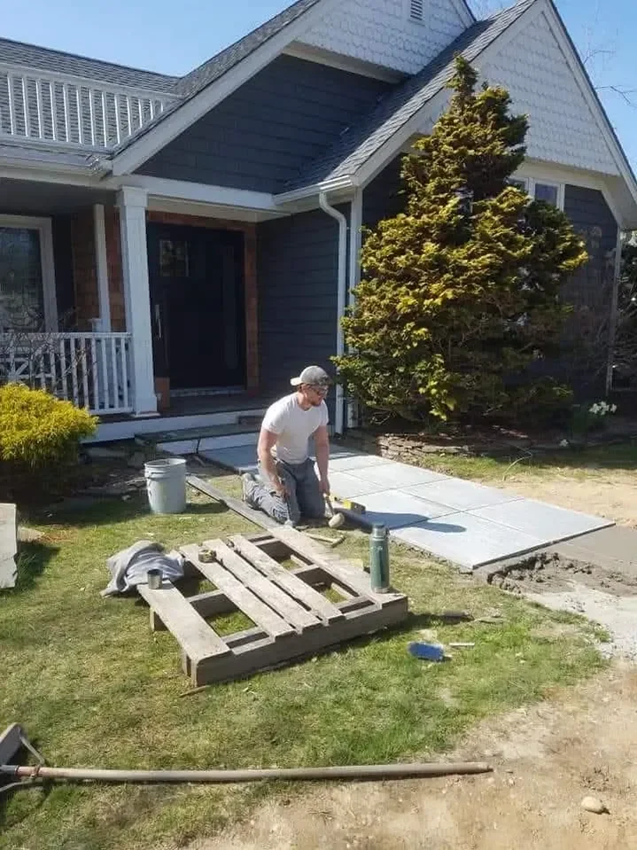 A construction worker kneels on the lawn installing large square pavers to create a new walkway leading up to the dark blue siding house entrance.