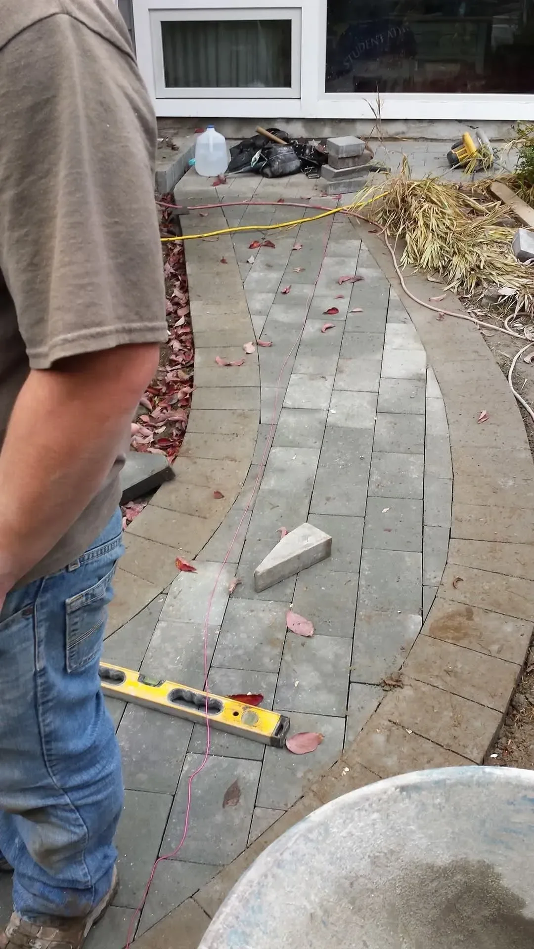 A worker stands on a partially completed curved paver walkway, surrounded by tools and construction materials next to a building foundation.