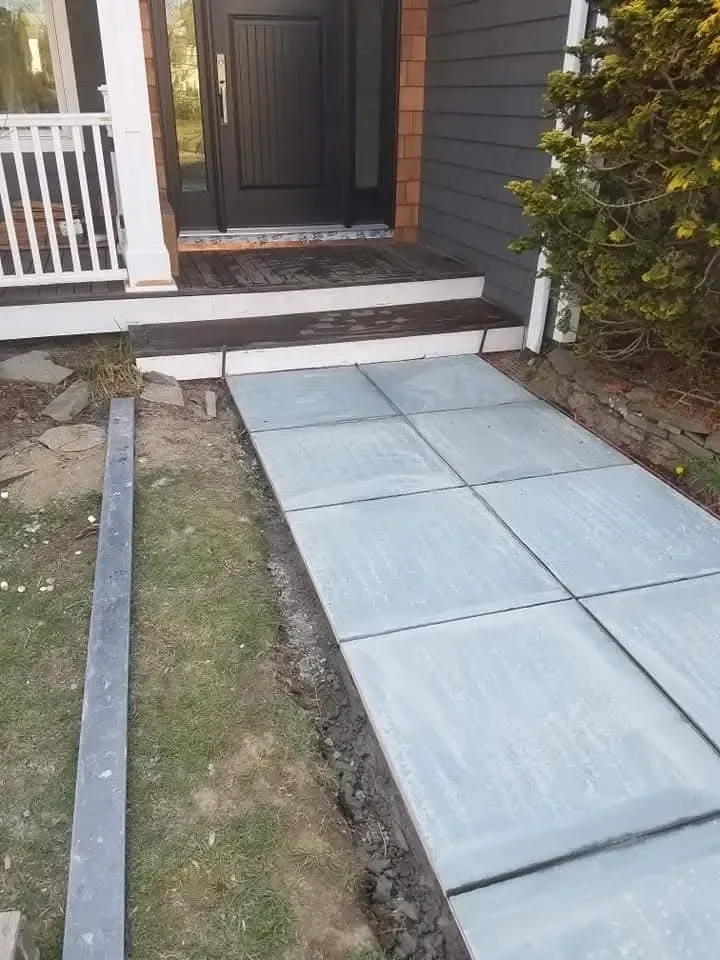 A newly poured concrete walkway leads to the dark front door and wooden steps of a residential home, surrounded by unfinished landscaping work.