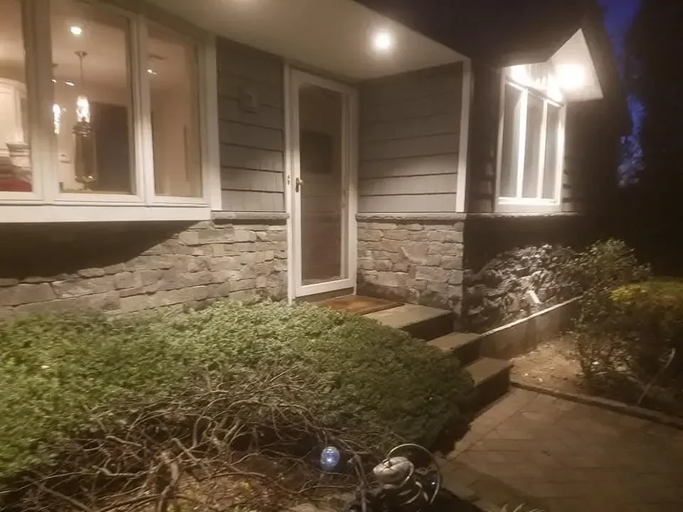A nighttime view of a home entrance featuring gray siding, stacked stone veneer, and illuminated landscaping leading to the front door.