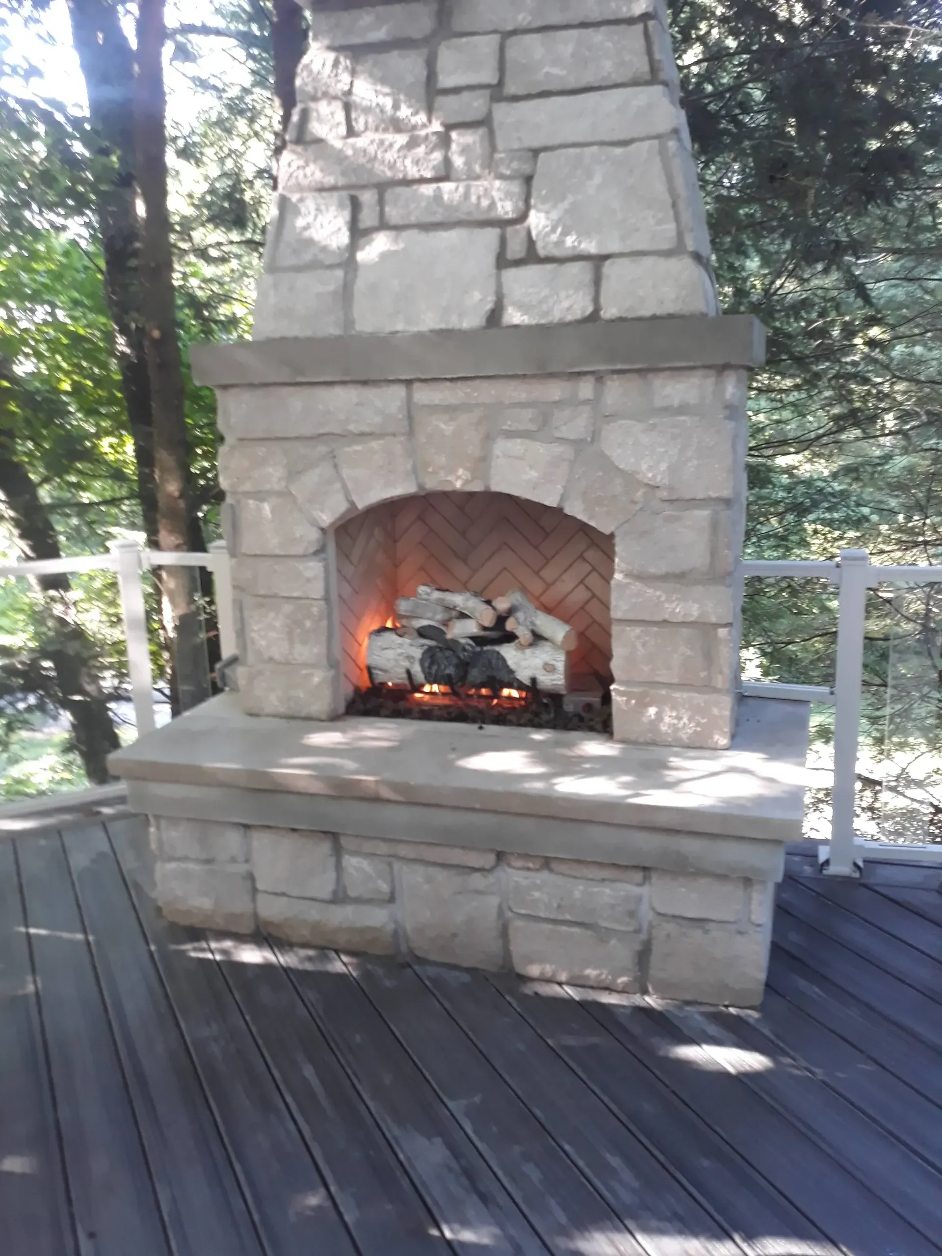 A large, rustic stone fireplace structure featuring a lit gas log insert is built upon a dark composite deck surrounded by lush green trees.