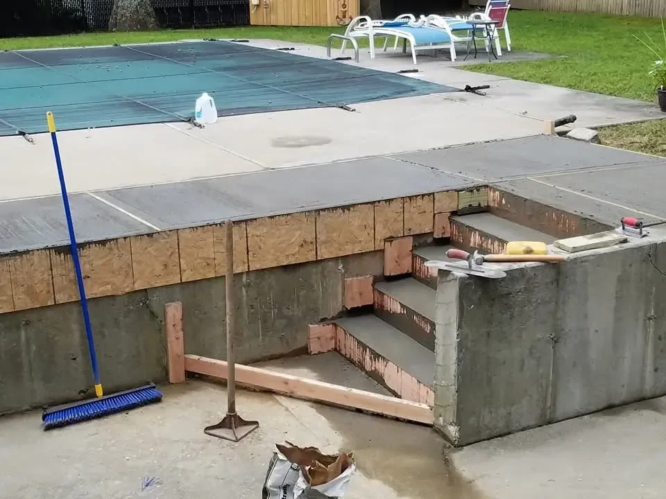 Freshly poured concrete steps are visible with wooden formwork and construction tools next to a covered swimming pool patio, indicating ongoing work.