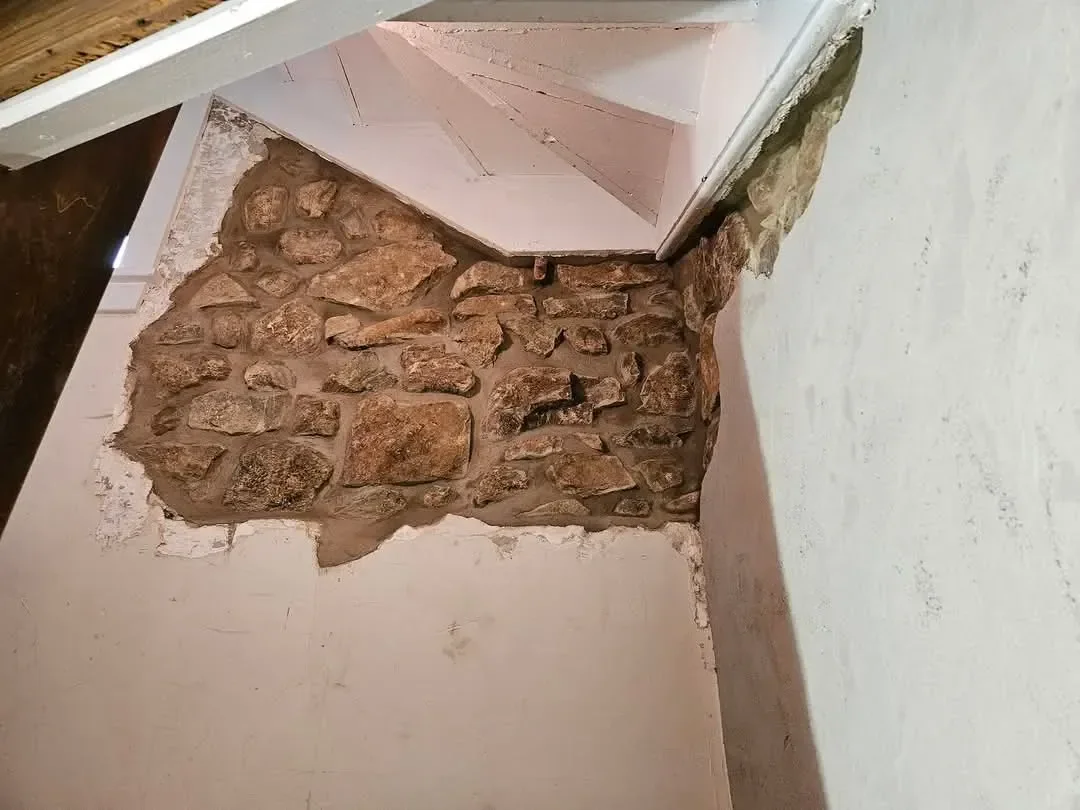 A section of plaster has been removed from an interior wall corner beneath a staircase, revealing the rough, historic stone masonry structure underneath.