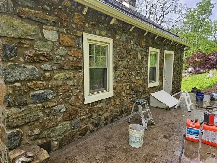 The rustic fieldstone exterior of a small building is shown with two windows and construction tools, including a ladder and buckets, suggesting ongoing cleaning or repair work.