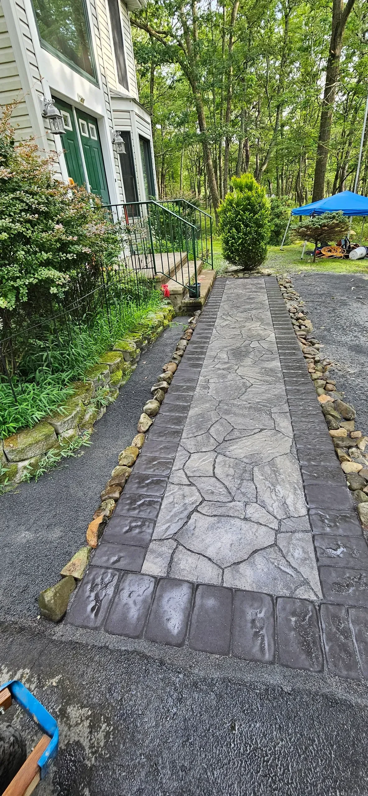 A newly installed residential walkway features a gray stamped concrete flagstone pattern bordered by dark pavers and natural river rocks leading to a house entrance surrounded by lush greenery.