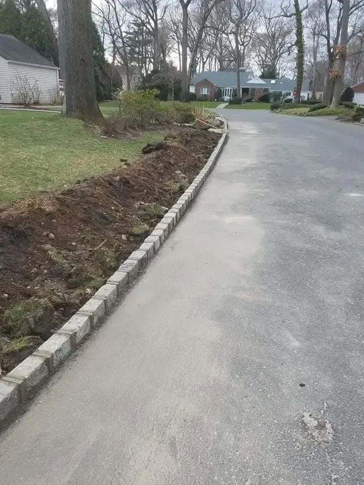 Newly installed stone edging separates a paved suburban driveway from a mound of excavated soil and grass, indicating ongoing landscaping work in a residential neighborhood.