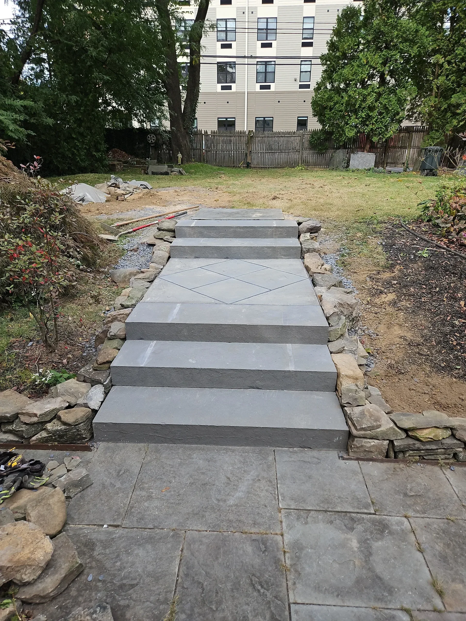 Newly installed gray stone steps, featuring a diamond pattern, ascend from a patio into a backyard flanked by natural stone edging, with construction materials visible nearby.