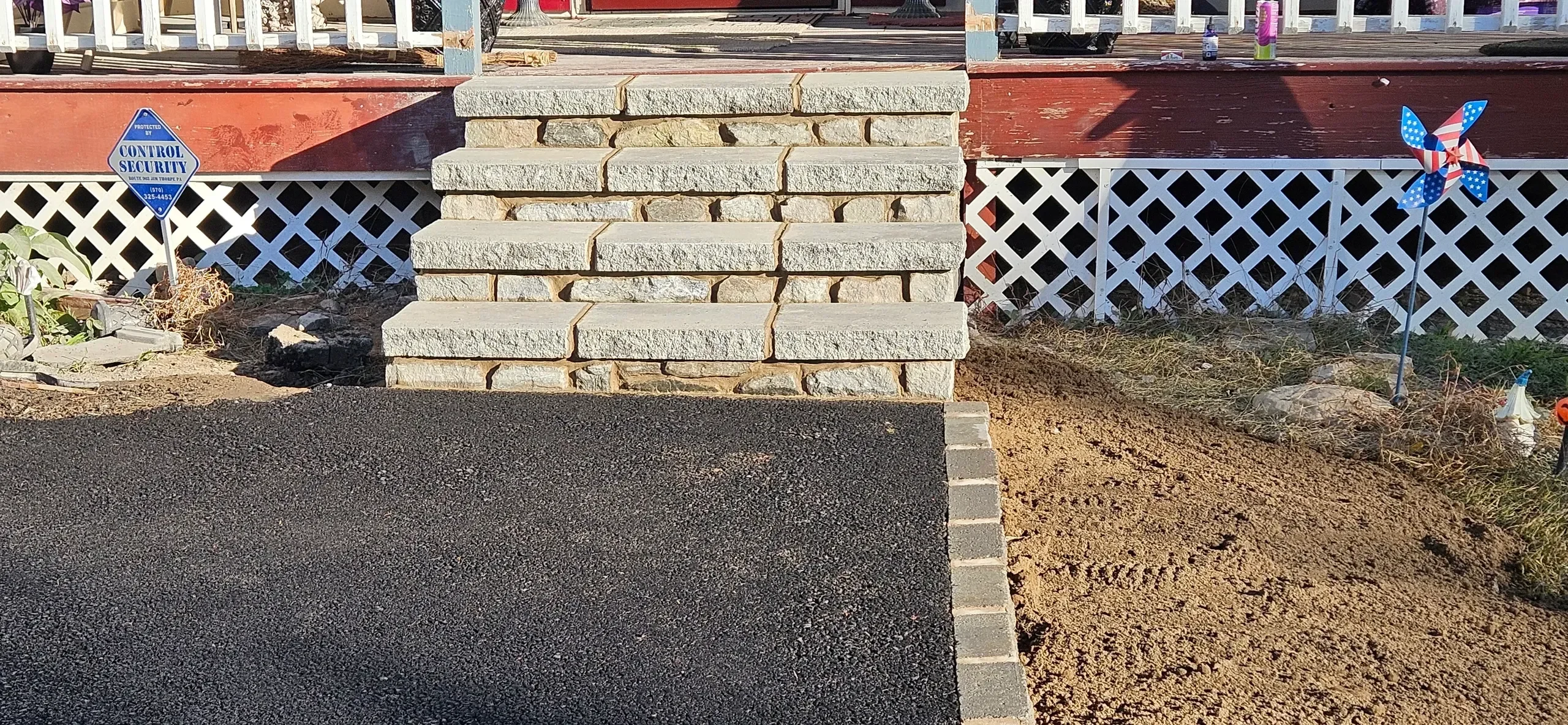 Newly installed stone steps connect a dark paved area to a residential porch featuring red siding and white lattice skirting, surrounded by disturbed earth.