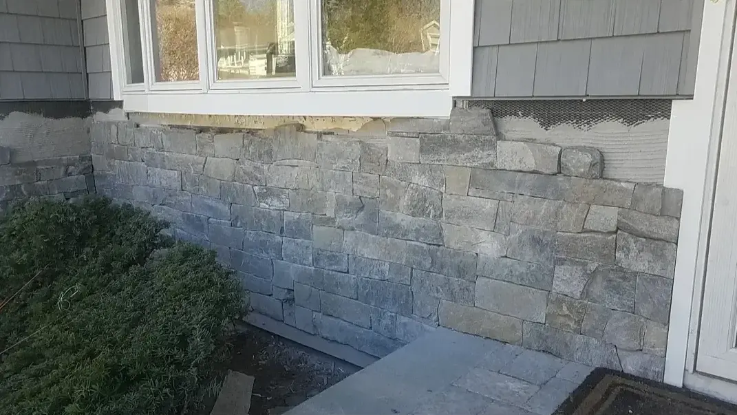 A residential foundation wall is being covered with gray, rough-cut stone veneer, showing exposed wire mesh and substrate indicating ongoing construction.