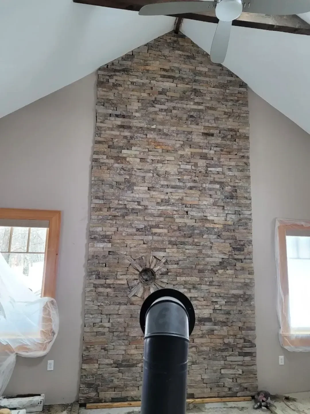 A tall interior wall featuring stacked stone veneer and a stovepipe installation extends up to a vaulted ceiling, with windows protected by plastic sheeting indicating ongoing construction.