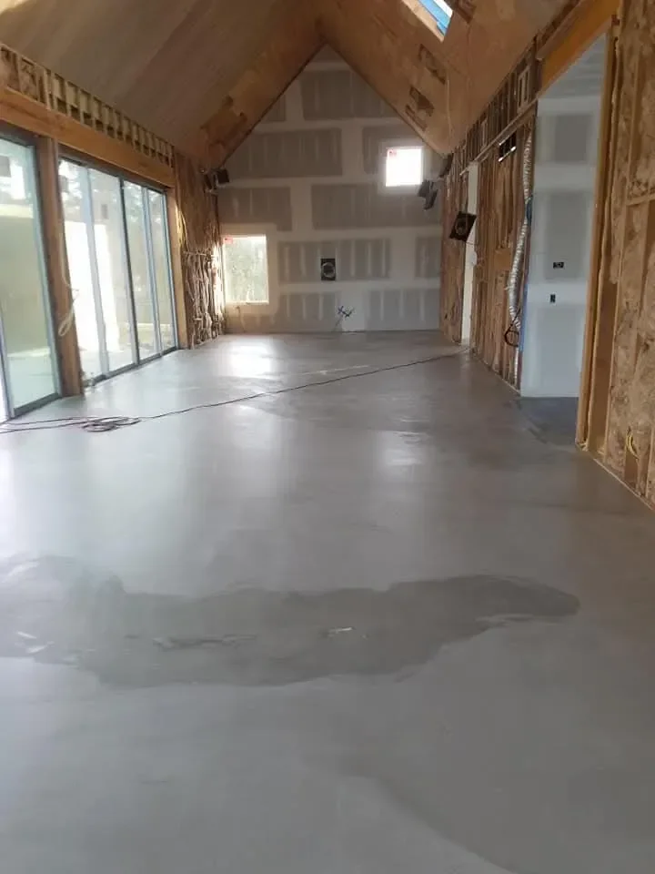 A large, vaulted room interior is under construction, featuring a newly poured concrete floor, exposed wood framing, and partially installed drywall.