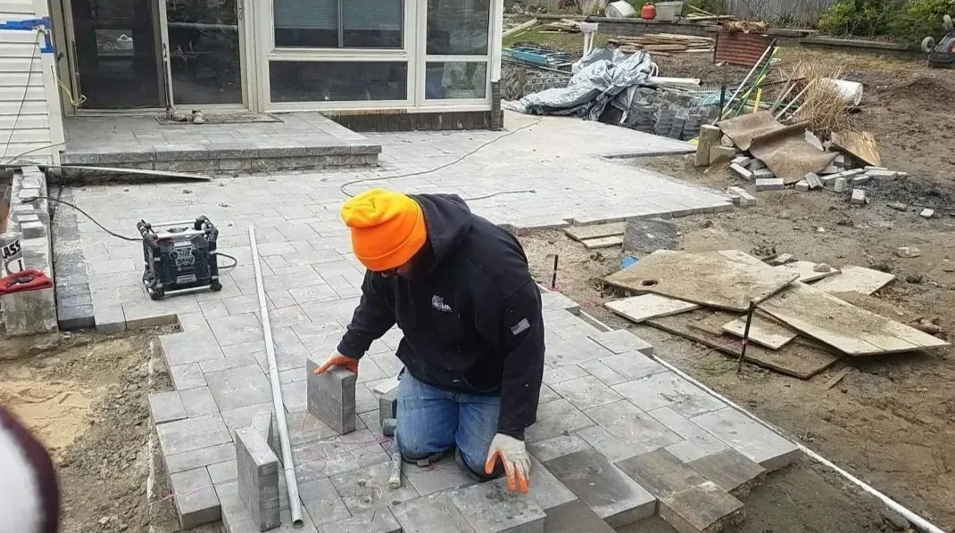 A construction worker kneels on the ground, carefully placing gray paving stones to create a new patio surface next to a residential building.