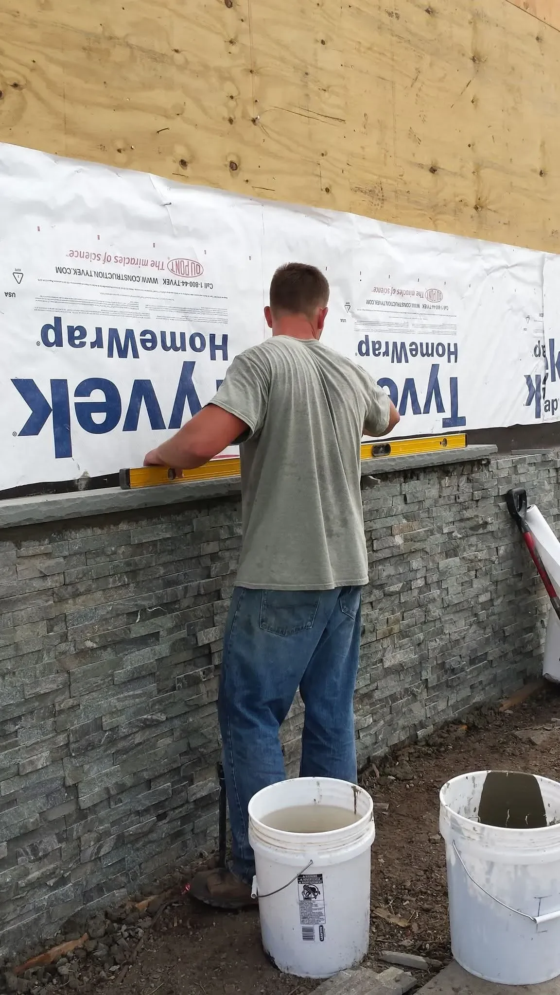 A construction worker uses a level to ensure proper placement of a capstone on a newly installed stacked stone veneer wall, with house wrap visible above.